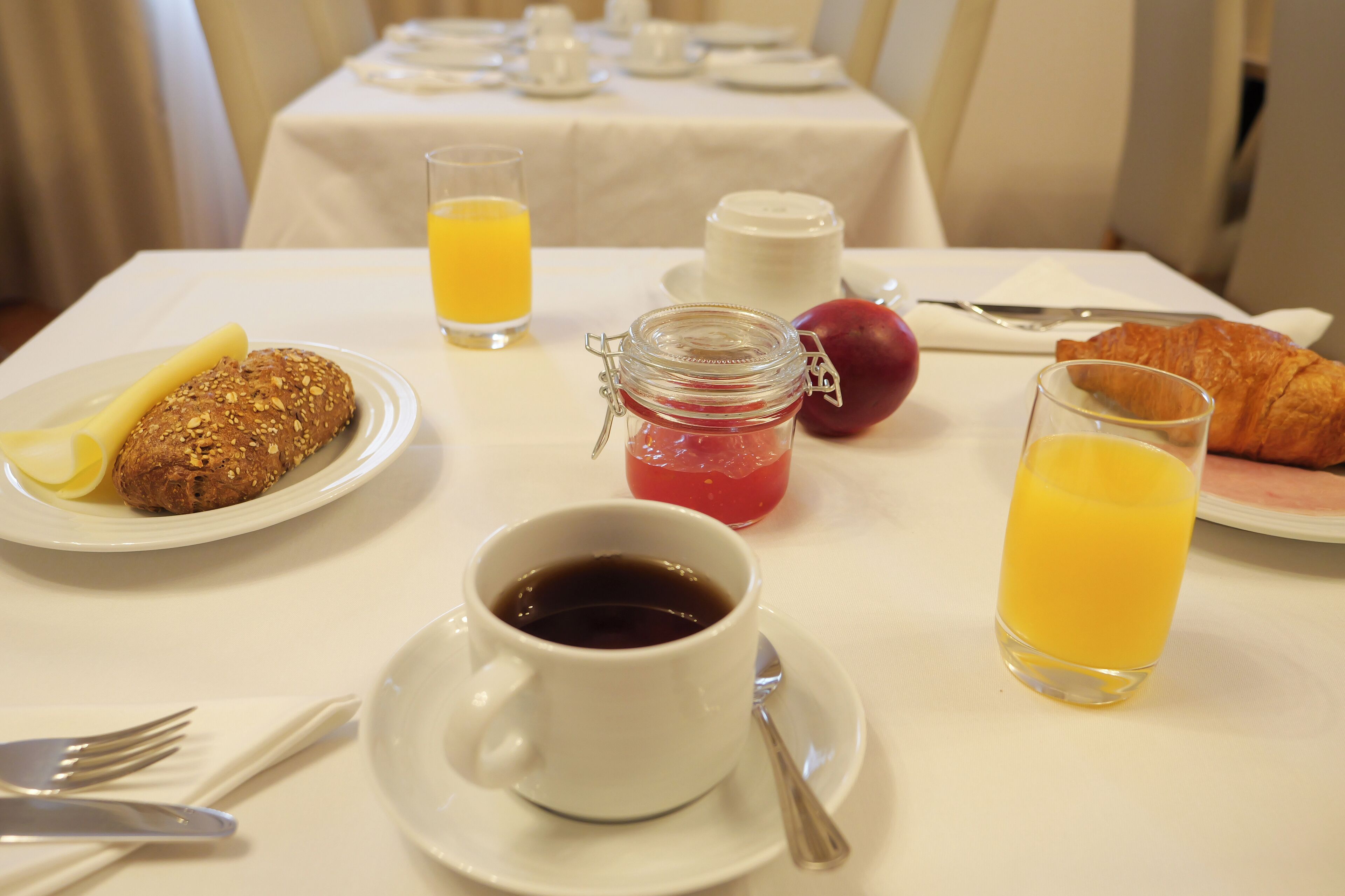 A breakfast table with croissants, a muffin, a glass of orange juice, a cup of coffee, a jar of jam, and a glass of milk.