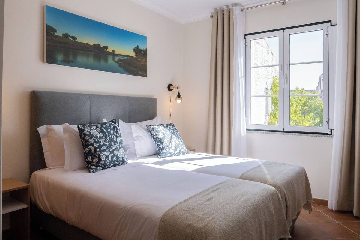 Ein Doppelbett mit grauem Kopfteil, Kissen in Weiß und Blau sowie eine beige Bettdecke. Ein Landschaftsbild hängt an der Wand, und es gibt ein Fenster mit Vorhängen.
