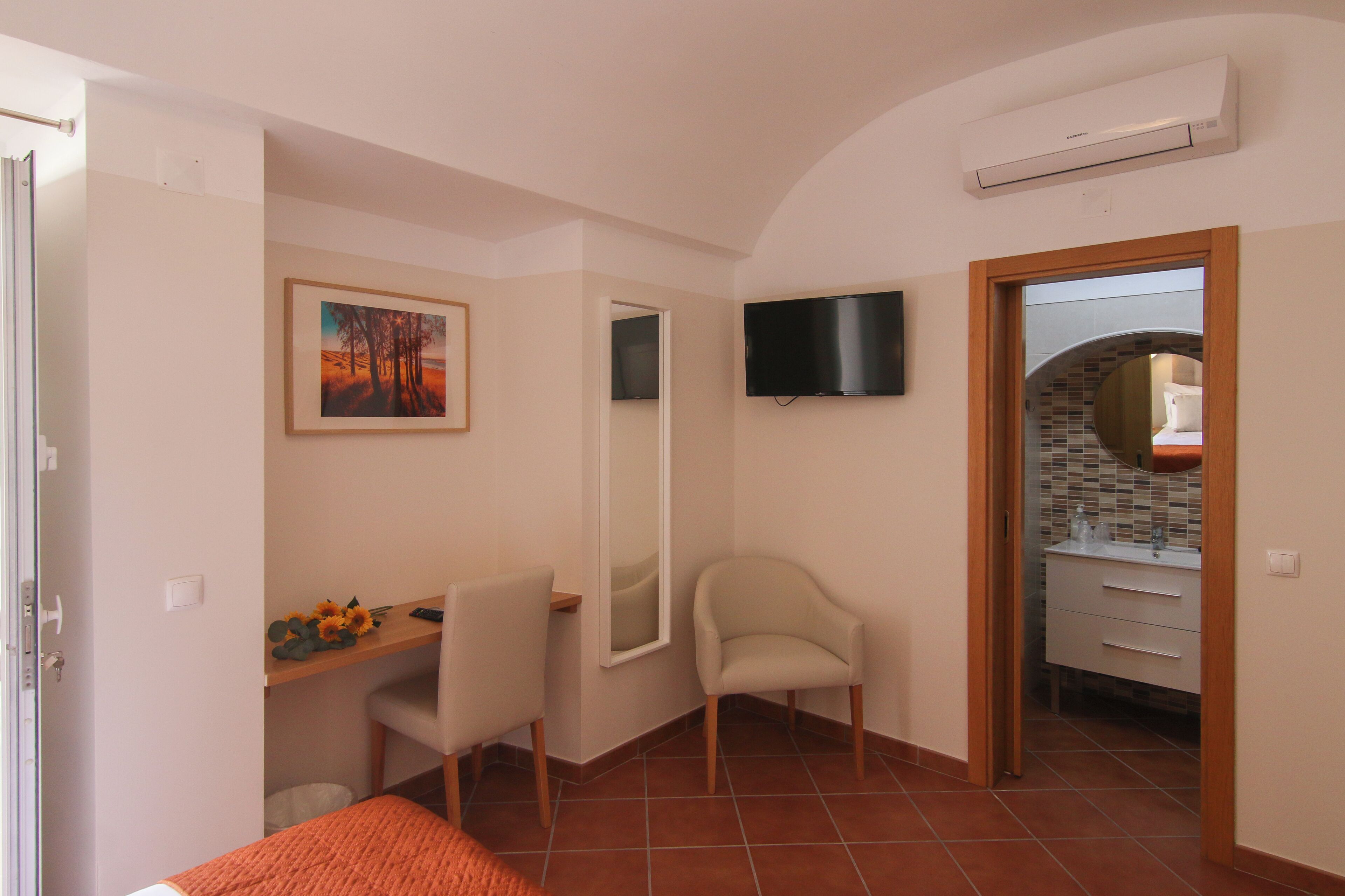A bedroom with a bed, a desk with a chair, a TV mounted on the wall, and a bathroom visible through an open door.