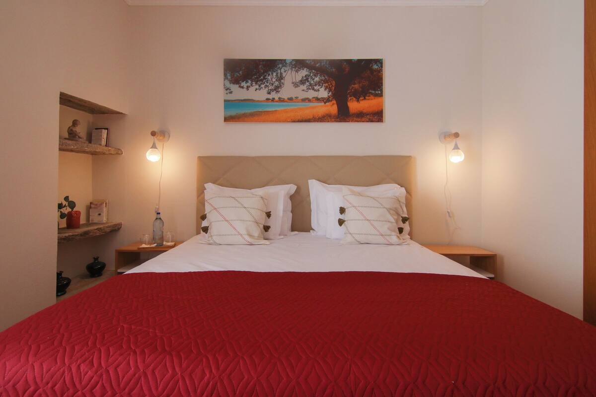 Ein Schlafzimmer mit einem großen Bett, zwei Nachttischen, einem Beistelltisch, einem Regal mit Dekoration und einem Gemälde an der Wand.