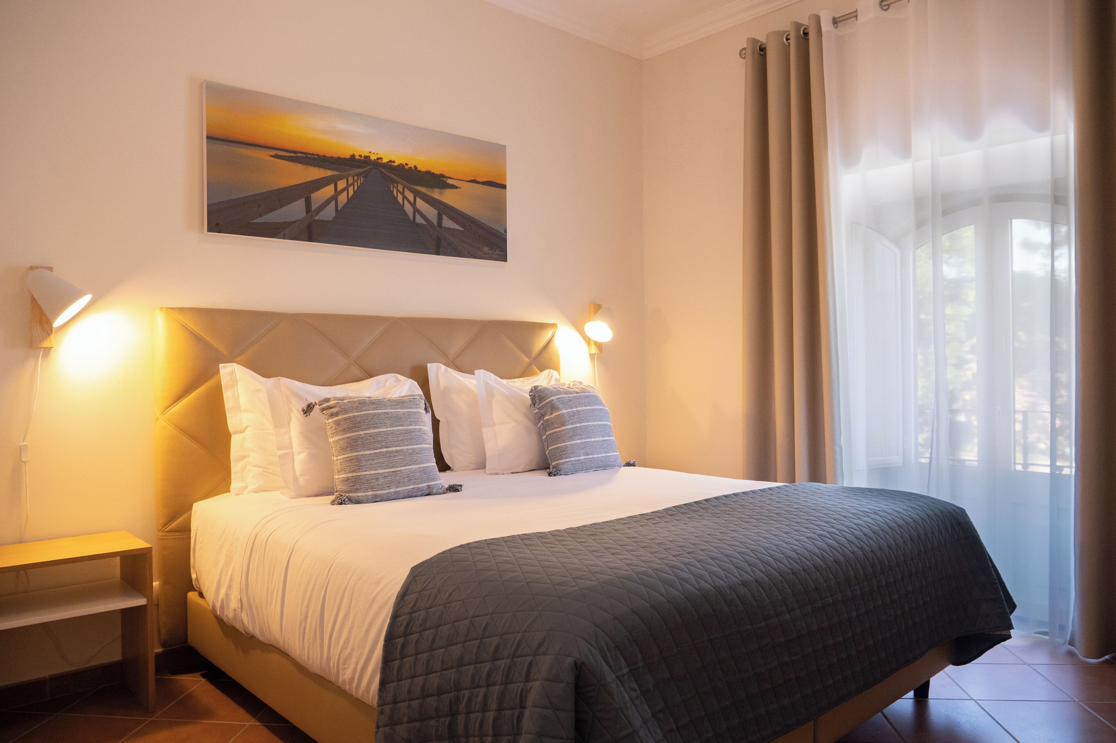 A neatly made bed with a quilted cover, two bedside lamps, a nightstand, and a painting of a sunset over a pier on the wall.