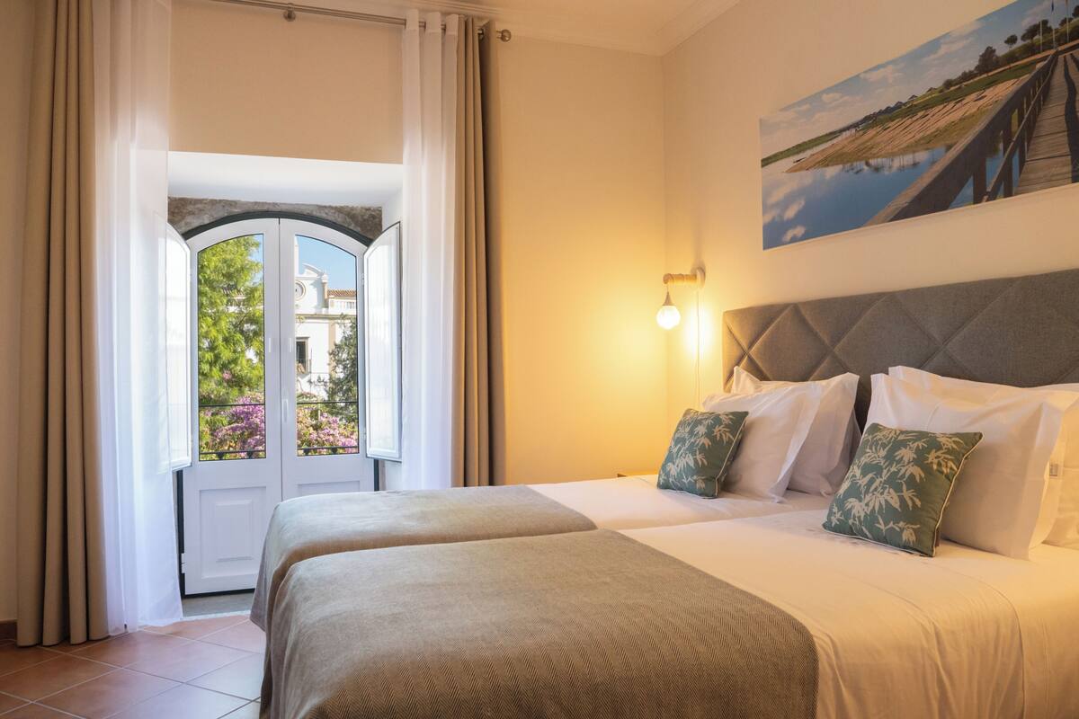 Ein Hotelzimmer mit einem Bett, zwei Kissen, einem Nachttisch, einem Gemälde und einem Fenster mit Vorhängen.