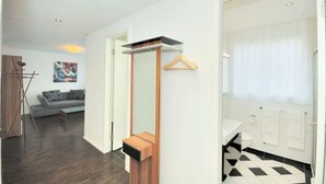 Quarto de hotel moderno com um espelho grande, um banco de madeira, um cabideiro suspenso, chão quadriculado em preto e branco e vista para uma sala de estar com sofá e uma pintura.