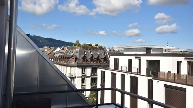 Vista de uma área residencial com edifícios, incluindo um prédio branco com persianas e varanda, sob um céu parcialmente nublado.