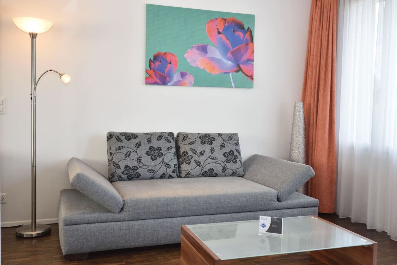 A living room with a grey sofa, a glass coffee table, a floor lamp, and a painting of flowers on the wall.
