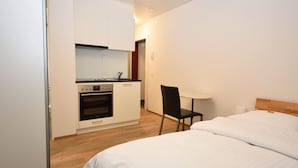 A compact room with a bed, a small kitchenette, and a chair.