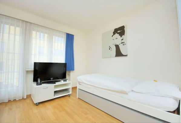 Een slaapkamer met een bed, een televisie op een witte standaard en een raam met dunne gordijnen.