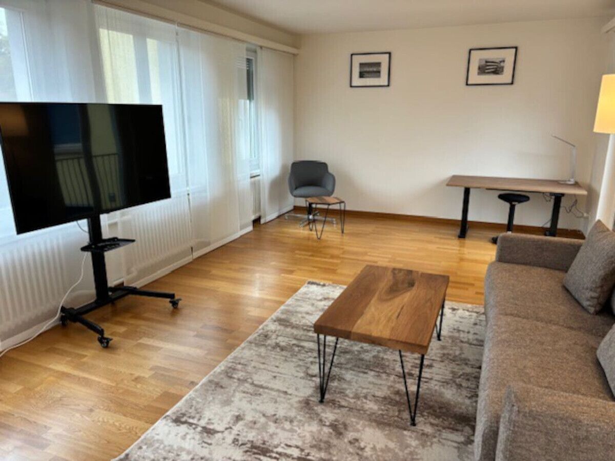 A modern living room with a flat-screen TV, a wooden coffee table, a grey armchair, a sofa, and a small table with a lamp.