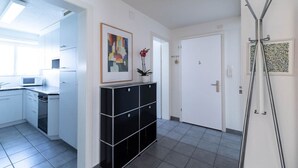 A modern kitchen with white cabinets, a microwave, and a stove. There is a black storage unit with drawers and a potted plant. A white door is visible in the background.