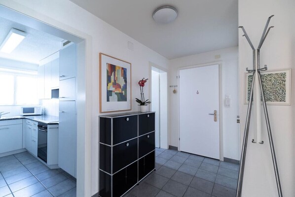 A modern kitchen with white cabinets, a microwave, and a stove. There is a black storage unit with drawers and a potted plant. A white door is visible in the background.