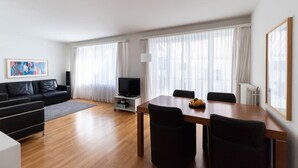 A modern living room with a black leather sofa, a wooden dining table with four chairs, a flat-screen TV on a stand, and large windows with sheer curtains.
