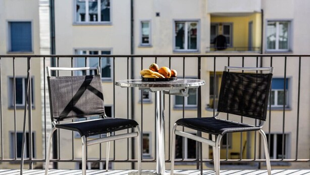 A balcony with two chairs and a small table, a bowl of fruit, and a view of a multi-story building.