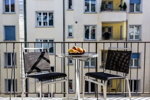 A balcony with two chairs and a small table, a bowl of fruit, and a view of a multi-story building.