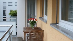 Un pequeño balcón con una mesa, sillas, un jarrón de flores y una ventana.