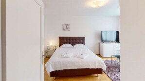 A neatly made bed with white linens, a brown headboard, and a flat-screen TV on a white cabinet. - Very spacious studio / city center (Parkview 4) (Zürich)