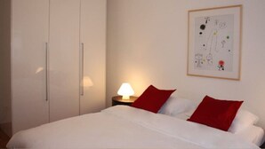 A bedroom with a bed, white walls, a wooden nightstand with a lamp, a framed abstract artwork, and a white wardrobe.