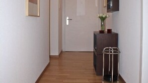 A hallway with wooden flooring, a door at the end, a small cabinet with a vase, and a mirror on the wall.