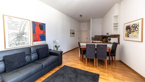 Een moderne woonkamer met een zwarte leren bank, een eethoek met een houten tafel en stoelen, en een keuken op de achtergrond.