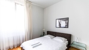 Een slaapkamer met een bed, een nachtkastje, een glazen tafel, een ingelijste foto aan de muur en een raam met gordijnen.