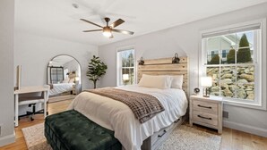 Quarto com cama, escrivaninha, cadeira, espelho, ventoinha no teto e janela com vista para pedras e árvores.