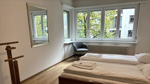 Quarto com duas camas, um radiador, uma cadeira e janela com vista para árvores e edifícios.