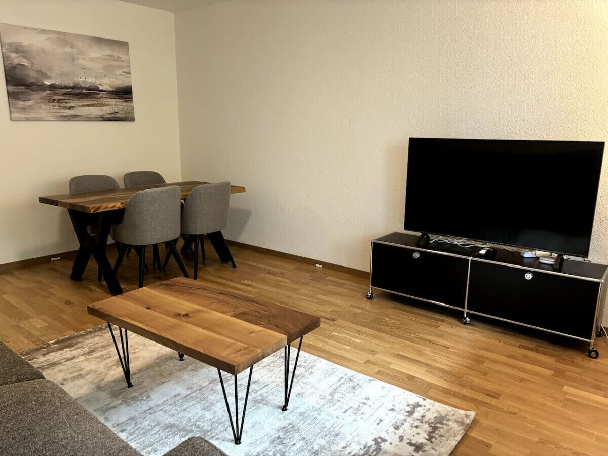 Un salon avec une table basse en bois, des fauteuils gris, un meuble TV noir et un tableau accroché au mur.