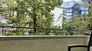 Uma varanda com uma mesa redonda e duas cadeiras, um muro de concreto e uma árvore com folhas verdes.