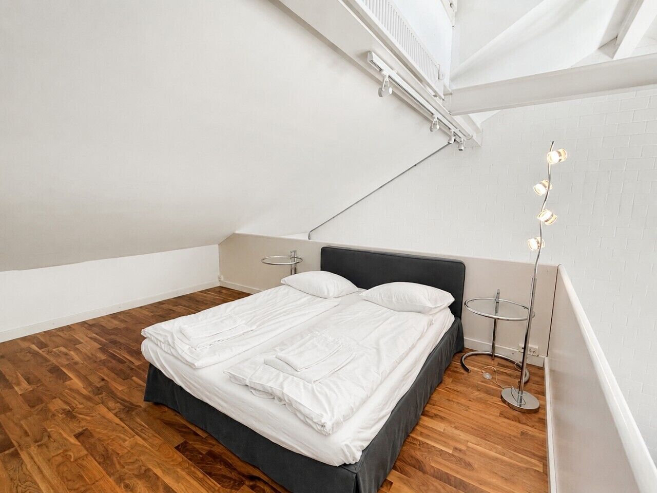 Cama bem arrumada em quarto com piso de madeira, cabeceira moderna e luminária de chão.