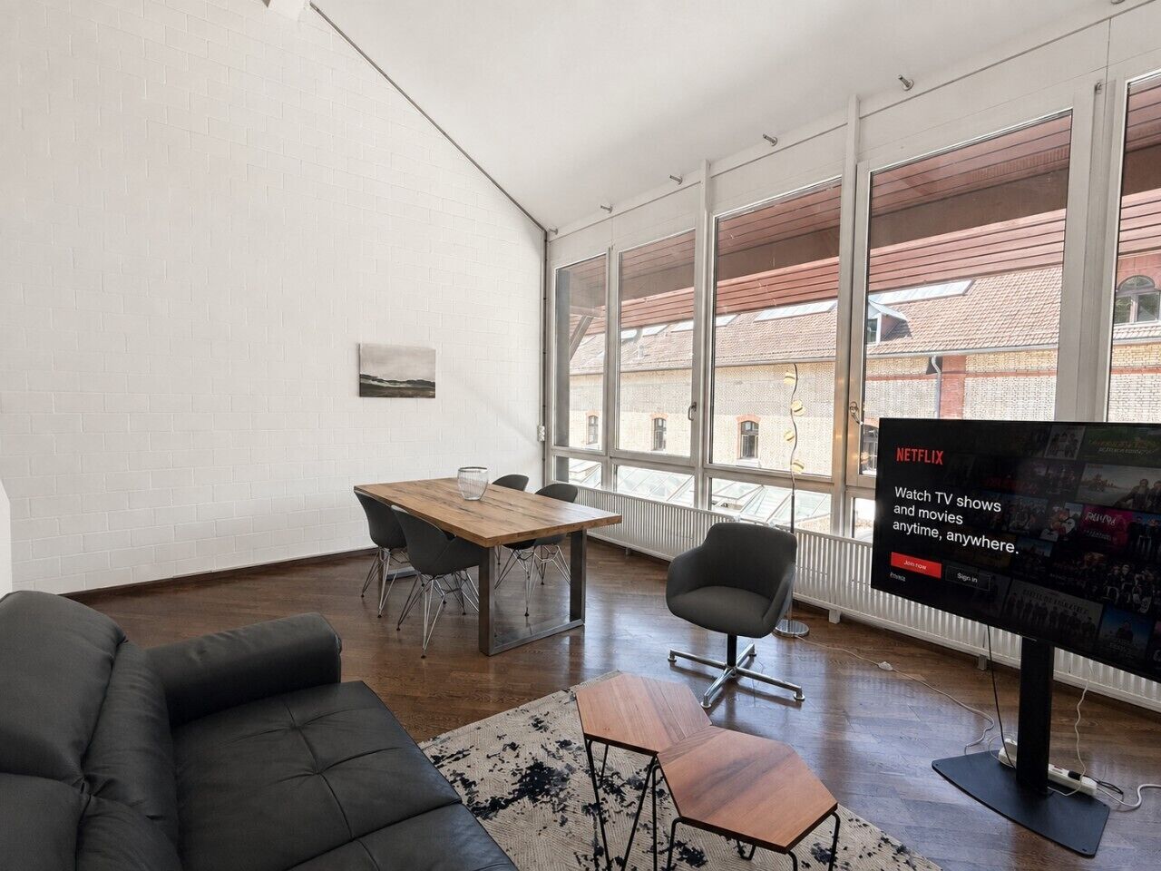 Ein modernes Wohnzimmer mit großem Fenster, einem Holztisch, einer schwarzen Couch und einem Flachbildfernseher, der eine Netflix-Werbung zeigt.