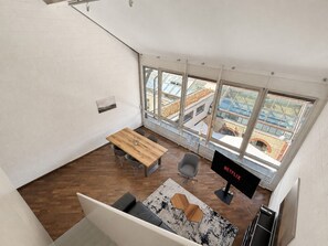 A high-ceiling room with a wooden floor, a dining table, chairs, a sofa, a TV, and large windows offering a view of buildings and a blue sky.