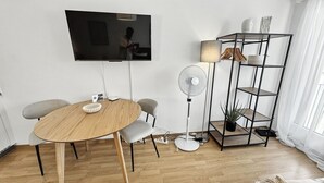 A modern living room with a wooden dining table, two grey chairs, a standing fan, a TV mounted on the wall, and a black metal shelving unit with a plant. - Amazing Central Studio - Crown 32 (Zürich)