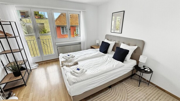 A hotel room with a large bed, a bedside table, a nightstand, a floor lamp, a window with a view of buildings, and a plant on a shelf. - Amazing Central Studio - Crown 32 (Zürich)