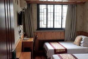 Quarto com escrivaninha de madeira, duas camas, janela com cortinas e televisão fixada na parede.