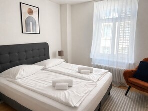 A double bed with white linens and two towels, a grey upholstered headboard, a side table with a lamp, and a window with sheer curtains.