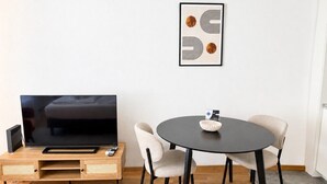 A modern living room with a flat-screen TV, a round dining table, two chairs, and a framed artwork on the wall.