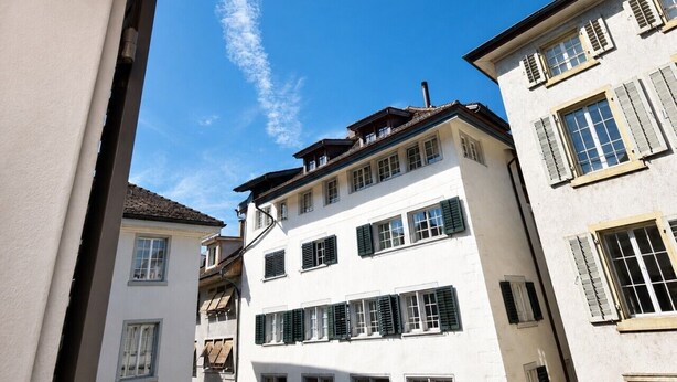 A multi-story building with a white facade and green shutters, featuring a balcony on the upper floor.