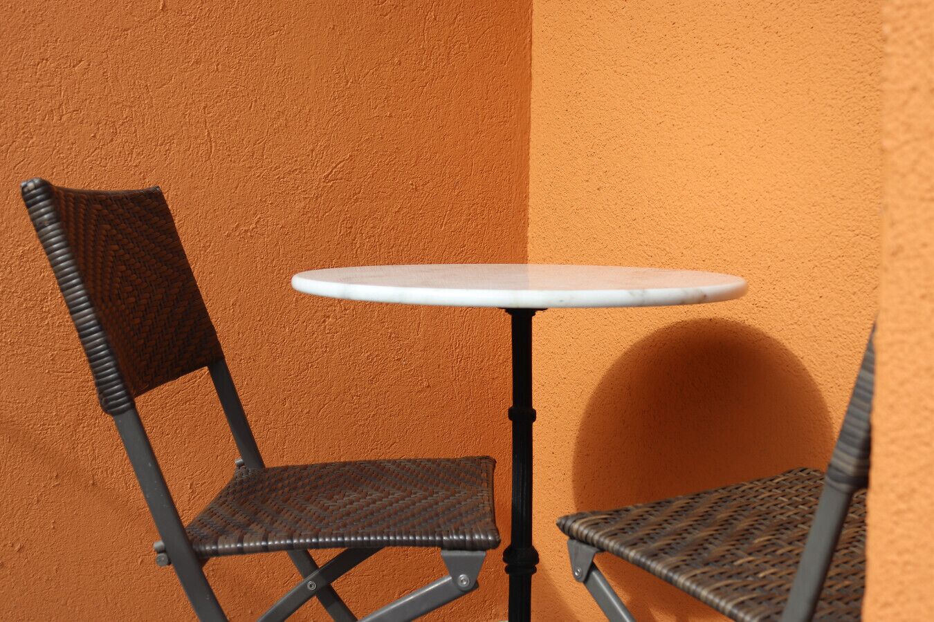 A small outdoor seating area with a round table and two wicker chairs against a textured orange wall.