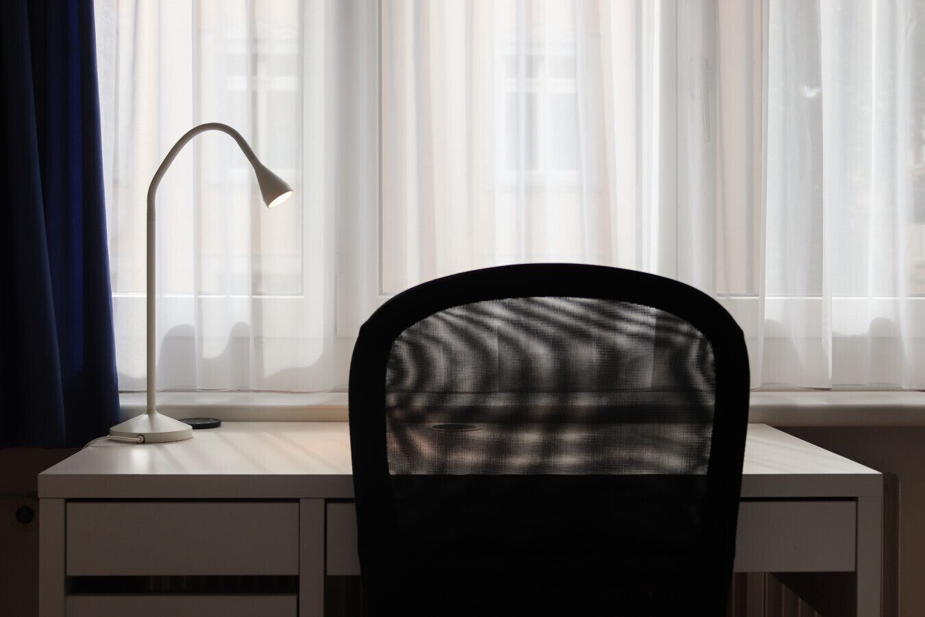 A desk with a lamp and a chair in front of a window with sheer curtains.
