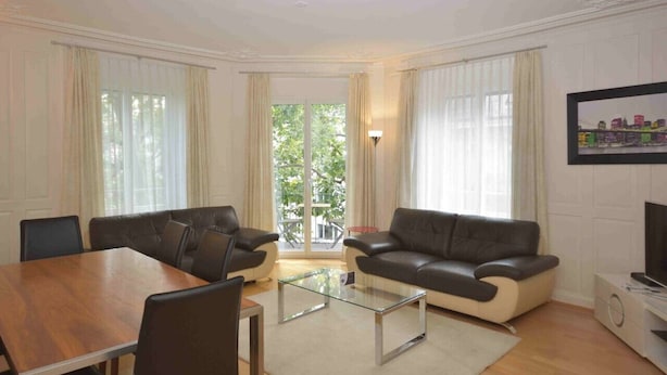 Una sala de estar con una mesa de comedor de madera, dos sofás de cuero negro, una mesa de centro de vidrio y un televisor sobre un mueble blanco.