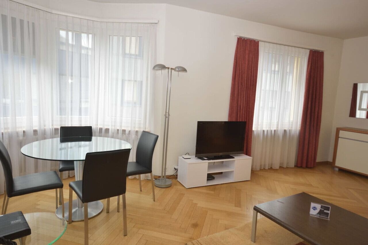 Un salon moderne avec une table ronde en verre, des chaises noires, un téléviseur à écran plat posé sur un meuble blanc, une lampe sur pied et une petite table sur laquelle repose une brochure.