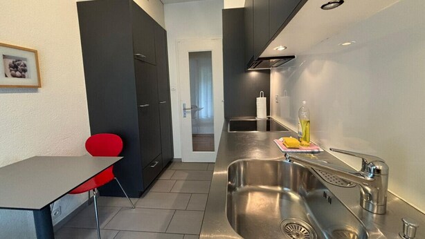 A modern kitchen with a stainless steel sink, a red chair, and a dark-colored cabinet. - Spacious 1BDR near Lake and Opera - Mill 3.11 (Zürich)