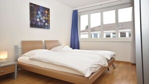 Un llit doble amb llençols blancs, una tauleta de nit amb una làmpada i una finestra gran amb cortines blaves.