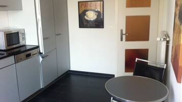 Une cuisine compacte équipée d’un four à micro-ondes, d’une petite table à manger et de chaises. Un tableau représentant une tasse de café est accroché au mur.