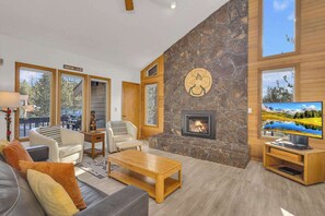 A modern living room with a stone fireplace, large windows, and a television. - Located in heart of Sunriver, complex pool access! (Sunriver)