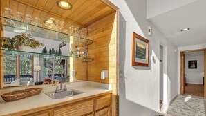 A kitchen with wooden cabinets, a glass shelf with wine glasses, a sink, and a window overlooking a deck. - Located in heart of Sunriver, complex pool access! (Sunriver)