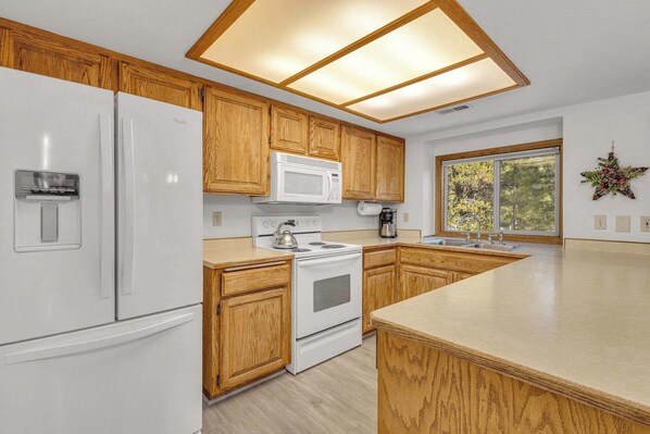 Une cuisine avec des meubles de rangement en bois, un réfrigérateur blanc, un four à micro-ondes, une cuisinière et une grande fenêtre.