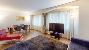 Een moderne woonkamer met een flatscreen televisie, een eettafel met stoelen, een rode bank en een groot raam met gordijnen.
