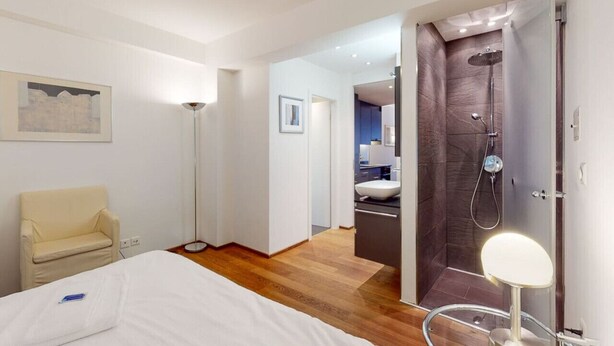 Een moderne hotelkamer met een bed, een bedlamp, een stoel, een vloerlamp, een douchegedeelte en een badkamer zichtbaar door een geopende deur.