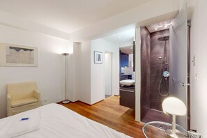 Une chambre d’hôtel moderne équipée d’un lit, d’une lampe de chevet, d’une chaise, d’un lampadaire, d’un espace douche et d’une salle de bain visible à travers une porte ouverte.