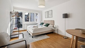 A modern bedroom with a bed, a bedside table, a floor lamp, a chair, a small table, and a view of the city through a window. - Studio in the heart of Zurich (Crown 24) (Zürich)